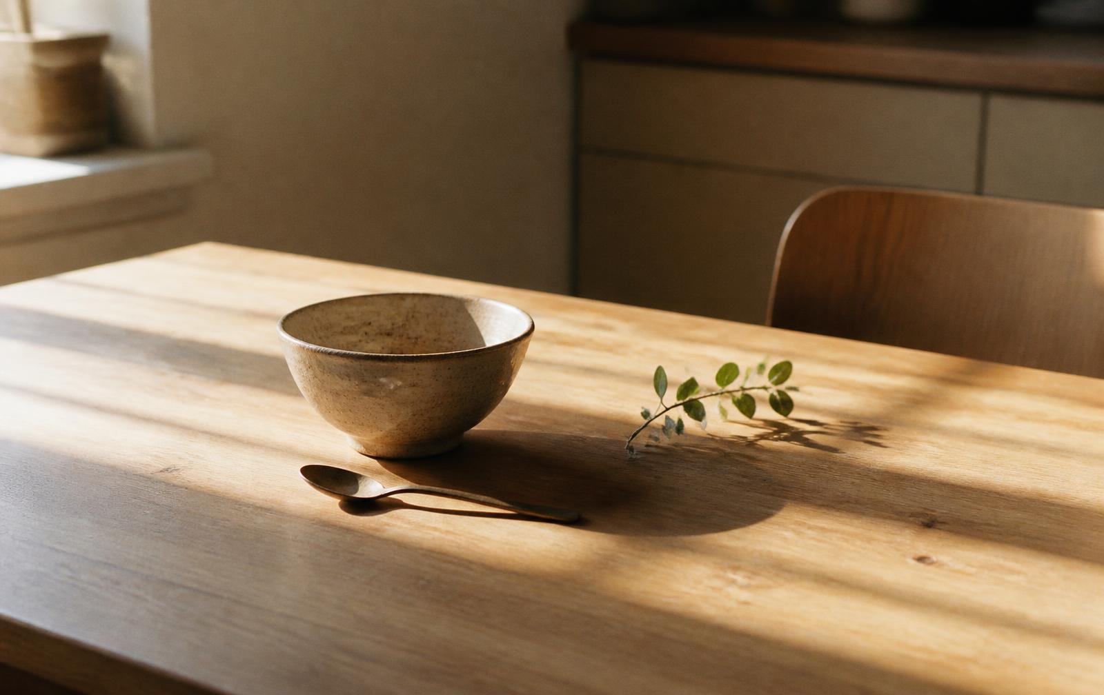 Morning sunlight across a bare wooden table with a bowl, a spoon and a sprig of greenery